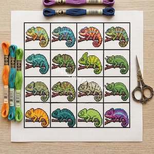May include: A cross-stitch pattern on white fabric featuring a grid of sixteen colorful chameleons perched on branches. The set includes three skeins of embroidery floss in orange, teal, and green, two purple and blue skeins, and metal scissors.
