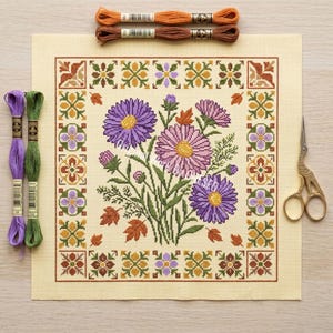 May include: A cross-stitch floral design featuring purple and pink flowers with yellow centres, surrounded by a decorative border. The design is on a cream-coloured fabric, with embroidery floss and gold scissors nearby.