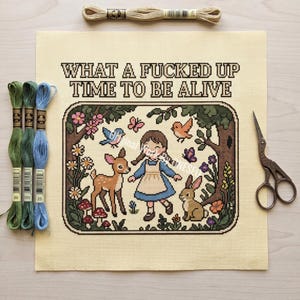 May include: A cross-stitch project on cream fabric featuring a girl, a deer, and a rabbit in a forest scene with the text WHAT A FUCKED UP TIME TO BE ALIVE. Includes three embroidery floss skeins in green and blue, and a pair of metal scissors.
