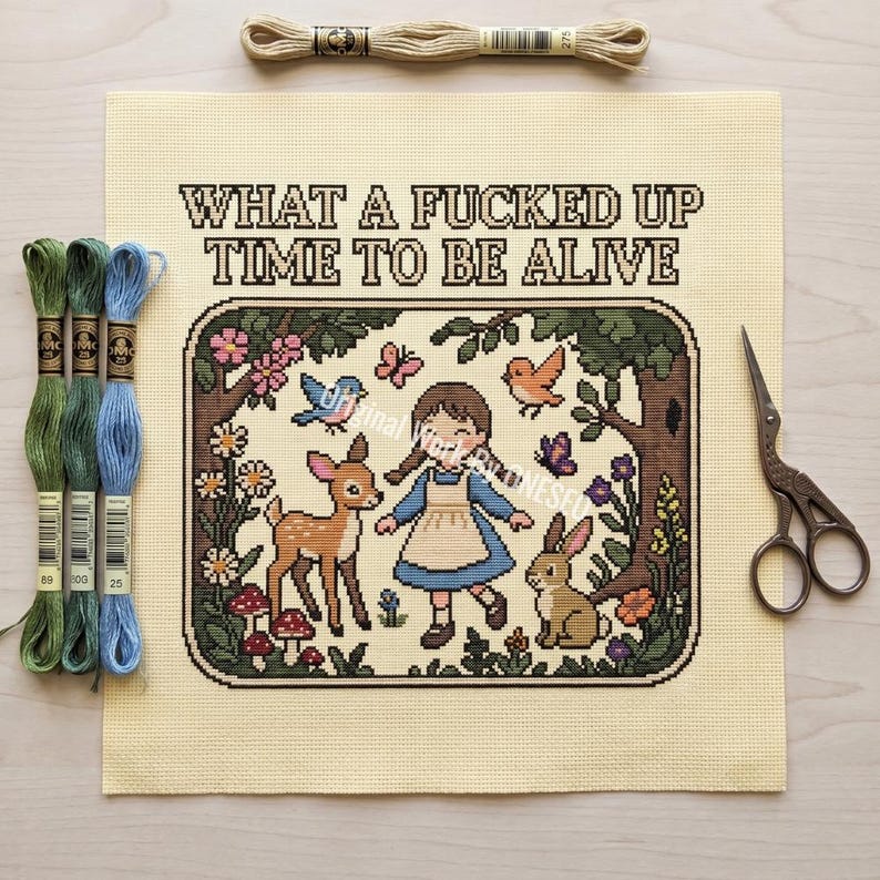 May include: A cross-stitch project on cream fabric featuring a girl, a deer, and a rabbit in a forest scene with the text WHAT A FUCKED UP TIME TO BE ALIVE. Includes three embroidery floss skeins in green and blue, and a pair of metal scissors.