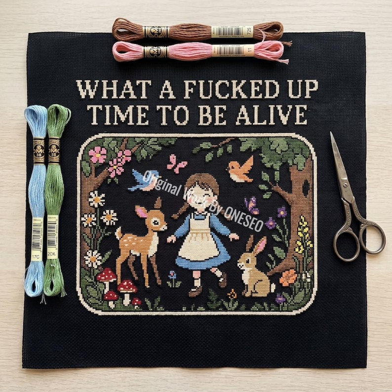 May include: A black cross-stitch fabric featuring the text WHAT A FUCKED UP TIME TO BE ALIVE above a scene of a person, a deer, and a rabbit in a forest. Includes four embroidery floss skeins in brown, pink, blue, and green, and metal scissors.