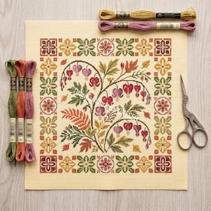 May include: A completed cross-stitch project featuring a floral design with heart-shaped flowers in shades of red and purple, surrounded by green leaves and a decorative border. Embroidery floss in various colors and small scissors are also present.