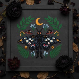 Könnte beinhalten: Ein gerahmtes Kreuzstichmuster mit einer stilisierten Motte mit detaillierten Flügeln, einem Halbmond und floralen Elementen. Die Motte ist schwarz mit farbenfrohen Flügeldetails. Das Design umfasst grüne Blätter, blaue Blumen und kleine rote und schwarze Insekten. Der Rahmen ist dunkelgrau.