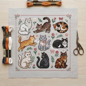 May include: A cross-stitch pattern featuring nine different cat designs in various poses and colors, surrounded by floral and bow accents. Embroidery floss and scissors are also visible, suggesting a crafting project.