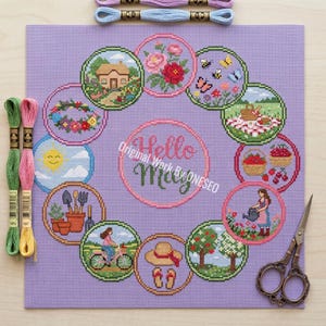 May include: A lilac-colored needlepoint canvas featuring a circular design with spring-themed illustrations. The design includes a house, flowers, bees, a picnic, and a woman watering flowers. The text "Hello May" is in the center. Embroidery floss and scissors are also visible.
