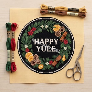 May include: A needlepoint kit featuring a black circular design with the words "HAPPY YULE" in white. The design includes a wreath with green leaves, red berries, orange slices, and cinnamon sticks. Embroidery floss and scissors are also present.