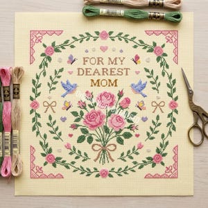 May include: A cross-stitch pattern on cream fabric featuring the text FOR MY DEAREST MOM, surrounded by a floral wreath of pink roses, green leaves, blue birds, butterflies, and small hearts, with embroidery floss and metal scissors nearby.