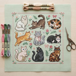 May include: A cross-stitch pattern featuring nine different cat designs in various poses and colors, surrounded by floral and bow accents. The pattern is on a light green fabric, with embroidery floss and scissors visible.