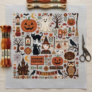 May include: A cross-stitch pattern on light gray fabric featuring Halloween motifs including pumpkins, a ghost, black cats, a witch, an owl, a haunted house, and signs reading PUMPKIN FESTIVAL, HALLOWEEN, and TRICK OR TREAT, with embroidery floss and scissors.