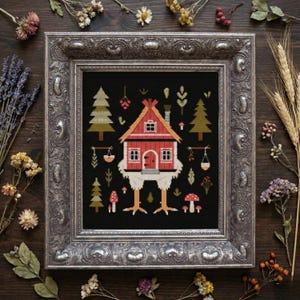 May include: Framed cross-stitch art depicting a quirky red house with chicken legs on a black backdrop. The house has a red roof and white trim, with green trees, mushrooms, and hanging plants. The frame is silver.