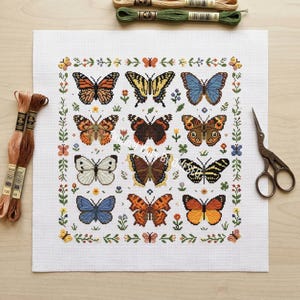 May include: A white Aida fabric square featuring a cross-stitch pattern of twelve distinct butterflies arranged in a grid, surrounded by a floral border. Two bundles of embroidery floss and a pair of vintage-style metal scissors are placed nearby.