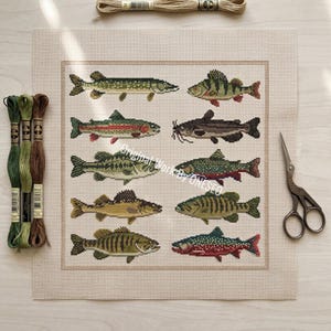 May include: A square piece of beige Aida fabric featuring a cross-stitch pattern of ten different fish arranged in two columns. Three skeins of embroidery floss in green and brown tones are on the left, and metal scissors are on the right.