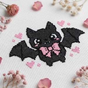 May include: A cross-stitch design featuring a black bat with pink accents. The bat has a pink bow and is surrounded by pink hearts. The background is white, with pink flower petals and dried flowers.