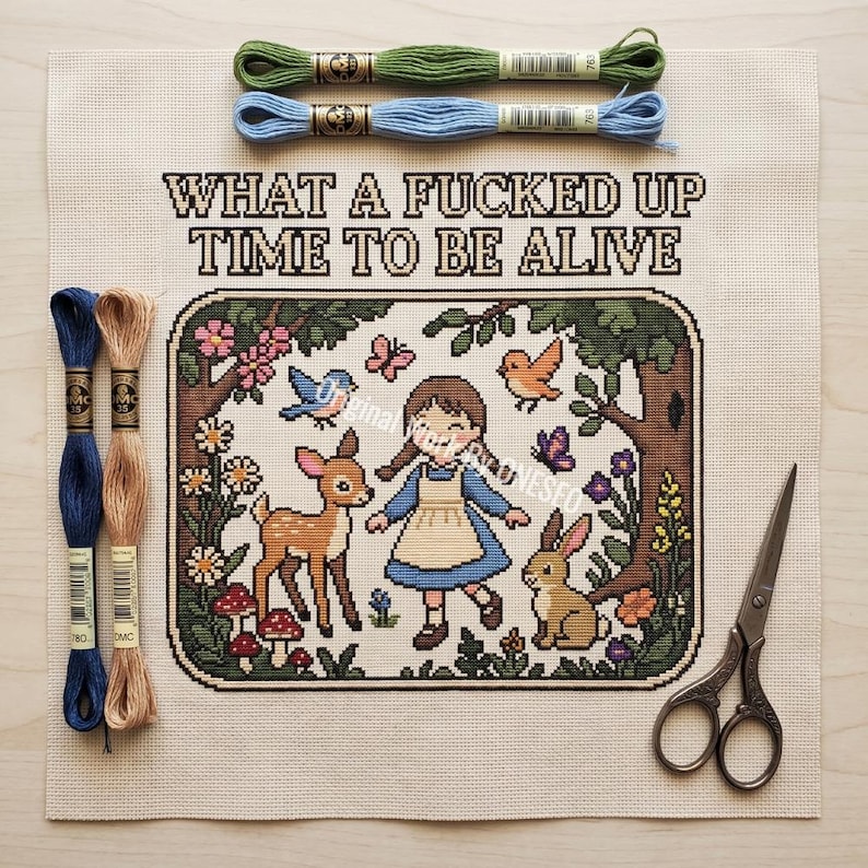 May include: A cross stitch pattern on beige fabric featuring a girl, a deer, and a rabbit in a forest with the text WHAT A FUCKED UP TIME TO BE ALIVE. Includes two green and blue embroidery floss skeins at the top, two blue and tan skeins, and scissors.