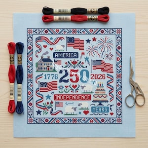 May include: A patriotic cross-stitch design on light blue fabric, featuring American flags, fireworks, and the text "America" and "Independence." Includes red, white, and blue embroidery floss and a pair of scissors. The design celebrates 250 years.