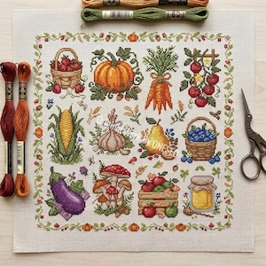 May include: A square cross-stitch fabric featuring a grid of harvest-themed motifs including a pumpkin, carrots, corn, mushrooms, eggplant, berries, and a jar of honey, surrounded by a decorative border, with embroidery floss and scissors.