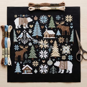 Op de afbeelding: Een zwart kruissteekproject met een winterbosscène met beren, elanden, bomen, sneeuwvlokken en hutten. Borduurgaren in verschillende kleuren en een schaar zijn rond het project geplaatst. Het ontwerp is in tinten wit, bruin, blauw en groen.