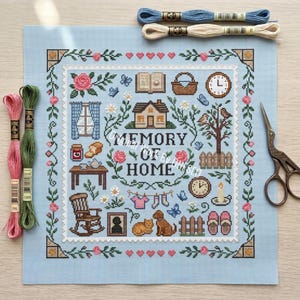 May include: A light blue cross-stitch fabric featuring the text "MEMORY OF HOME" surrounded by icons of a house, tree, rocking chair, and household items. Includes pink, green, and blue embroidery floss skeins and vintage-style metal scissors.