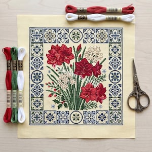 May include: A cross-stitch floral design featuring red amaryllis flowers and white blossoms, framed by a blue and green geometric border. Embroidery floss in red, green, and white, along with scissors, are arranged around the canvas.