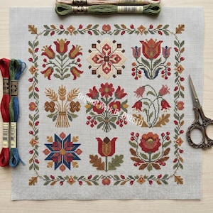 May include: Cross-stitch embroidery with a floral folk art design. The piece features a light gray background with a border of leaves and flowers. The design includes red, orange, yellow, and blue floral patterns. Embroidery floss and scissors are also visible.