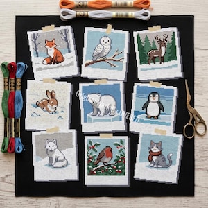 May include: A collection of nine cross-stitch designs featuring winter animals, including a fox, owl, deer, rabbit, polar bear, penguin, cat, robin, and another cat. The designs are displayed as polaroid-style images on a black fabric, with embroidery floss and scissors.