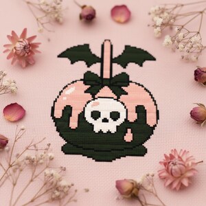 Può includere: Design a punto croce di una mela caramellata con un teschio, ali di pipistrello e un fiocco. La mela è verde scuro con glassa rosa gocciolante. Il teschio è bianco. Lo sfondo è rosa, con fiori e petali secchi.