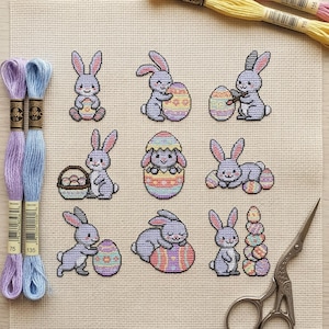 Könnte beinhalten: Kreuzstich-Osterdesign mit neun Hasen, Eiern und Körben. Die Hasen sind in verschiedenen Posen, einige halten Eier, andere Körbe, und einer ist in einem Ei. Das Design verwendet Pastellfarben und befindet sich auf einem beigen Stoff.