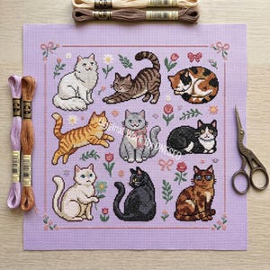 May include: A cross-stitch pattern featuring nine different cat designs in various poses and colors, surrounded by floral and bow accents. The design is on a lavender fabric, with embroidery floss and scissors visible.