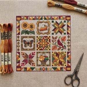 May include: A cross-stitch sampler featuring autumn-themed squares. The design includes sunflowers, a slice of pie, a butterfly, a flower, a cardinal, and a basket of fruit. The color palette includes shades of orange, yellow, green, and brown. Embroidery floss and scissors are also visible.
