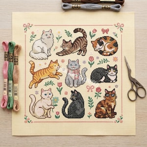 May include: A cross-stitch pattern featuring nine different cat designs in various poses and colors, surrounded by floral and bow motifs. The needlepoint is on a light beige fabric, with embroidery floss and scissors nearby.