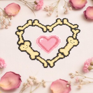 May include: Cross-stitch design featuring a heart-shaped frame of yellow bones with smiling faces, surrounding a pink pixelated heart. The design is on white fabric, with pink flower petals and sprigs in the background.