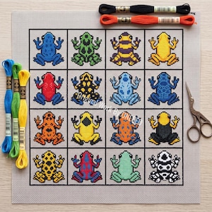 May include: A cross-stitch pattern on light gray fabric featuring a grid of sixteen colorful frogs in various patterns. Included are three skeins of embroidery floss in blue, green, and yellow, two loose skeins in black and orange, and metal scissors.