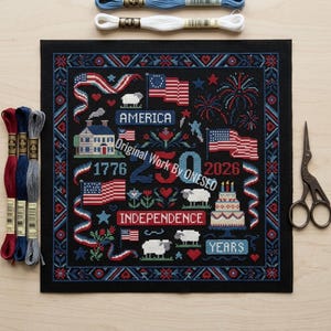 May include: A black cross-stitch design featuring patriotic symbols like American flags, fireworks, and sheep. The design includes the words "America," "Independence," and "Years." Embroidery floss and scissors are also visible.