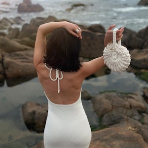 Könnte beinhalten: Eine Frau in einem weißen Häkelkleid mit Trägern steht an einem felsigen Strand und hält eine weiße Häkeltasche.