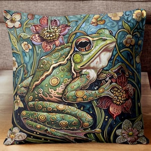 May include: Decorative square pillow featuring a detailed illustration of a green frog amidst flowers and foliage. The frog has large eyes and is surrounded by red, white, and yellow flowers against a teal background. The pillow has a soft, textured appearance.