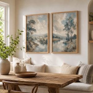 May include: Two vertical framed wall art prints featuring a landscape painting of a winding river flowing through a valley with trees and a cloudy sky. The artwork uses a muted color palette of soft blues, creams, and earthy browns in wooden frames.