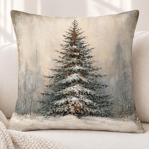 May include: A square throw pillow featuring a printed illustration of a single evergreen tree covered in snow. The scene depicts a winter forest landscape with muted beige, grey, and white tones, showcasing a detailed pine tree centered on the fabric.