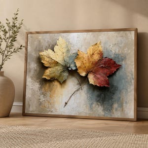 May include: A rectangular framed wall art piece featuring a textured painting of two large maple leaves. The left leaf displays muted green and yellow tones, while the right leaf transitions from yellow to deep red. The background is a neutral beige.