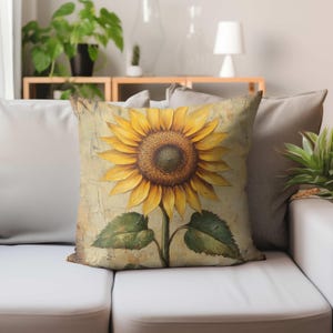 May include: A square throw pillow featuring a centered, large yellow sunflower with a dark brown textured center. The flower has long, pointed petals and two green leaves at the base. The background is a light beige with a cracked, aged texture.