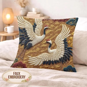 Puede incluir: Cojín cuadrado decorativo con un diseño bordado de dos grullas blancas, negras y azules sobre un fondo dorado. El cojín lleva la etiqueta "FAUX EMBROIDERY". El diseño evoca el arte tradicional.