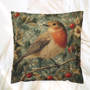 Könnte beinhalten: Ein quadratisches Zierkissen mit einer detaillierten Illustration eines Rotkehlchens auf einem Ast. Der Vogel hat eine rot-orange Brust und braune Flügel. Der Hintergrund ist in Salbeigrün gehalten, mit weißen Blüten, grünen Blättern und roten Beeren.
