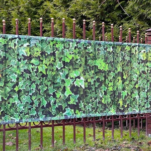 May include: A green ivy printed privacy screen attached to a metal fence. The screen is made of a lightweight material and is designed to provide privacy and shade.