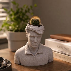 May include: A light grey, textured bust-style planter shaped like a person wearing a collared shirt with a zip detail. The top of the head is open, containing a small, round, green cactus with fine spines. The sculpture has a matte, stone-like finish.