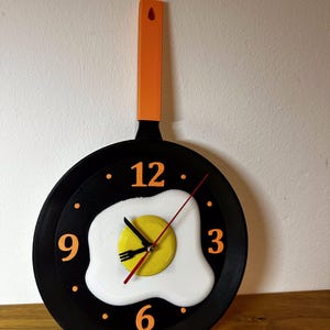 Peut inclure: Une horloge murale fantaisie en forme de poêle à frire avec un motif d'œuf au plat. Le cadran de l'horloge est noir avec des chiffres et des points orange. L'œuf a un jaune d'œuf jaune et un blanc d'œuf blanc. La poignée est orange.
