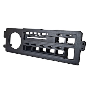 May include: A black plastic car radio console with various openings for controls and a circular opening on the left side. The console has a rectangular shape with multiple rectangular and square openings.