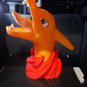 May include: A 3D-printed orange Charizard figure with red flames at the base. The figure has blue eyes and white teeth. The build volume is 256 x 256 x 256mm.