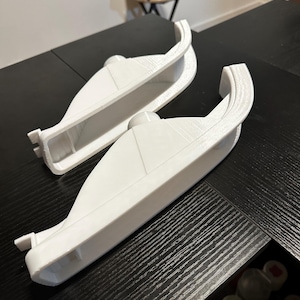 Puede incluir: Dos objetos blancos en forma de barco, con asas redondeadas y un botón central, se muestran sobre una superficie oscura. Los objetos tienen un aspecto liso y limpio, lo que sugiere un diseño moderno. Probablemente son componentes de un producto más grande.