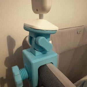 May include: A light blue plastic clamp mount attached to the side of a grey cot. The mount features a large adjustment knob on the side and a smaller knob for tilting the white baby monitor camera unit that is secured to the top of the device.