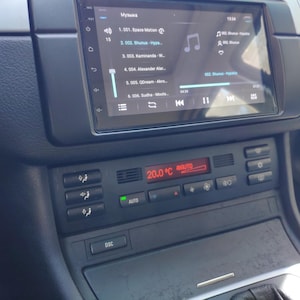 May include: A car dashboard featuring a digital display with a music playlist and climate control settings. The screen shows song titles and a temperature of 20.0°C. Air conditioning and other car function buttons are also visible.