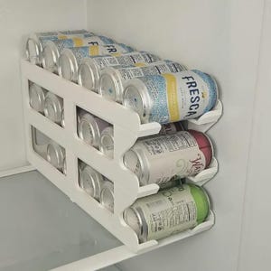 以下が含まれることがあります： 複数のアルミ製ソーダ缶を収納した、白色のプラスチック製多段式飲料缶オーガナイザーラック。最上段には青と黄色のラベルが付いたFresca缶が並び、下段には他の様々な飲料缶が積み重ねられた状態で収納されています。